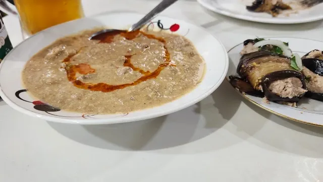 קערת מרק סמיך עם קו שמן אדום, לצד צלחת גלילי חציל וכוס בירה
