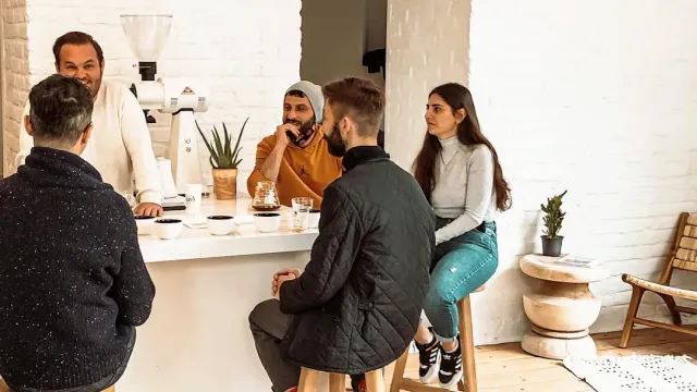 אנשים יושבים על שרפרפים סביב דלפק לבן עם כוסות קפה בחדר בהיר