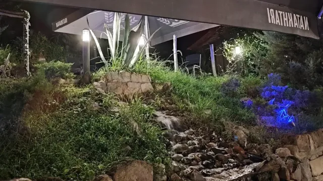 גינת סלעים מוארת בלילה עם פלג מים קטן וסוכך מעל שולחנות ברקע