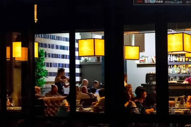 מבט מהרחוב אל חלל מואר בפנסי תקרה צהובים, עם סועדים ליד השולחנות ובר