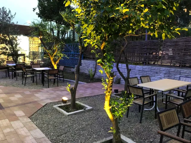 חצר מרוצפת בשעת ערב עם שולחנות, עצים מוארים מלמטה וגדר נצרים