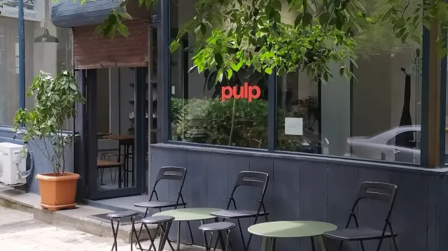 חזית כהה עם כיתוב אדום על החלון, כיסאות מתקפלים ושולחנות עגולים על המדרכה