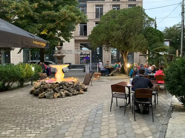 שולחנות ברחבה מרוצפת עם מזרקה מוארת וסועדים תחת עצים בשעת ערב