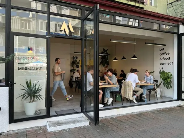 חזית זכוכית פתוחה לרחוב עם אורחים סביב שולחנות עץ וצמחים בפנים