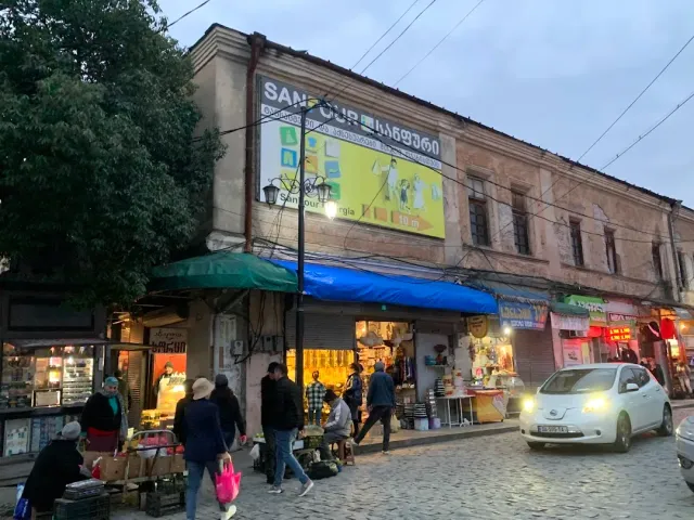 דוכני רוכלים תחת סוככים כחולים וירוקים ברחוב מרוצף, לפני בניין ישן עם שלט פרסומת